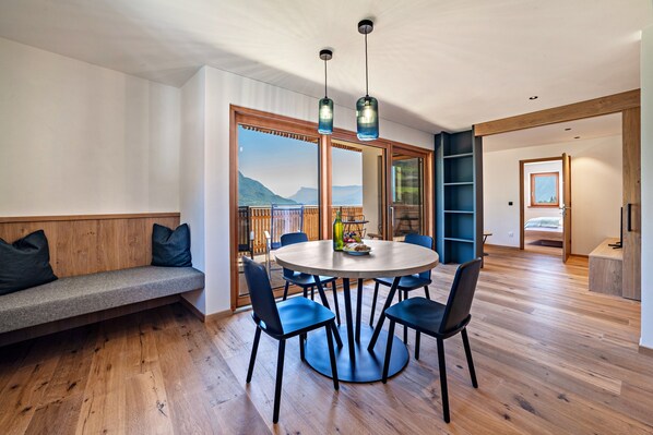Un espace repas moderne avec une table ronde, des chaises bleues, un banc en bois et une vue sur les montagnes grâce à de grandes fenêtres.