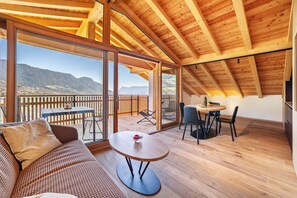 Ein geräumiges Zimmer mit Holzbalkendecke, großen Schiebeglasfenstern, einem runden Tisch und einer Couch.