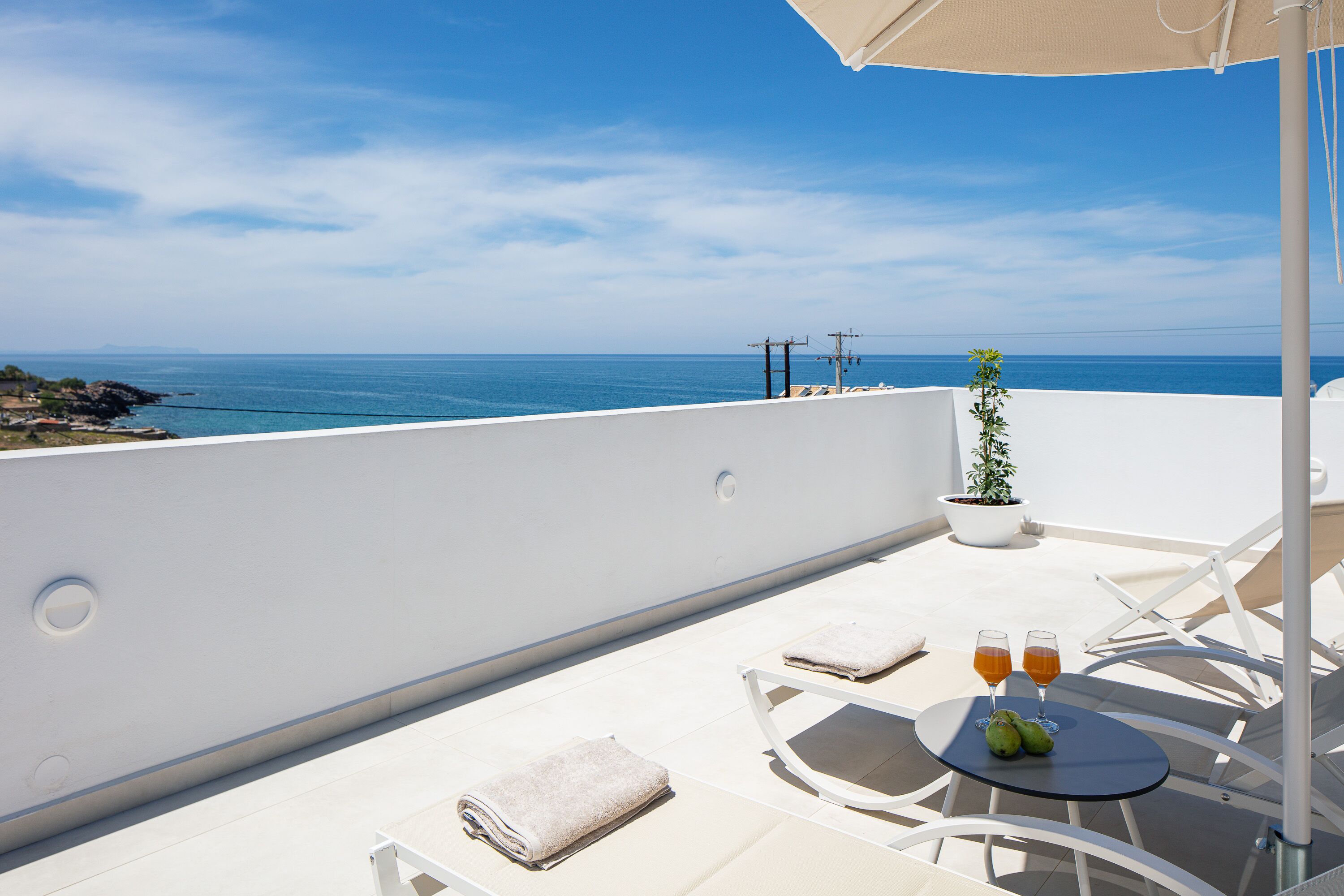 Un balcon avec vue sur la mer, une table avec des verres et des fruits, et une plante en pot.