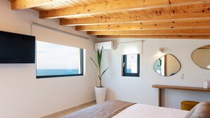 Ein Schlafzimmer mit einem großen Bett, Holzbalken an der Decke, einem Blumentopf, einem Flachbildfernseher und Meerblick durch ein Fenster.