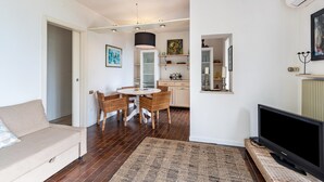 A living room with a sofa, a dining table with chairs, a flat-screen TV, and a kitchen area with cabinets and a hanging light fixture. - Apartment 'Villa Verde' with Lake View, Shared Pool and Air Conditioning (Torri del Benaco)