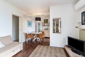 A living room with a sofa, a dining table with chairs, a flat-screen TV, and a kitchen area with cabinets and a hanging light fixture.