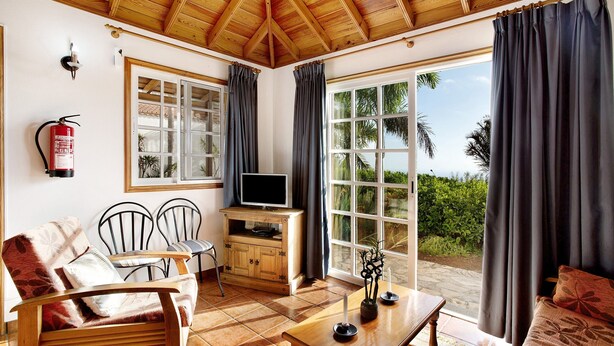 Una habitación con techo de madera, un televisor sobre un mueble de madera, una mesa de comedor con sillas y un ventanal con cortinas.