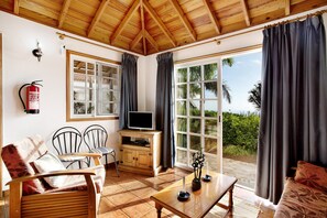 Une pièce avec un plafond en bois, un téléviseur posé sur un meuble en bois, une table à manger avec des chaises, et une grande fenêtre avec des rideaux.
