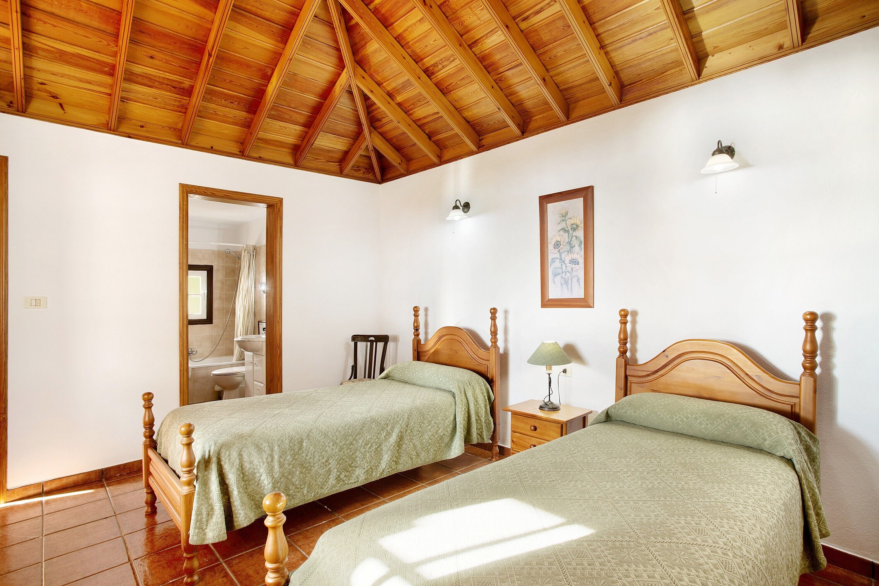 Een slaapkamer met twee houten bedden, een houten plafond, een nachtkastje met een lamp, en een badkamer die door een geopende deur zichtbaar is.