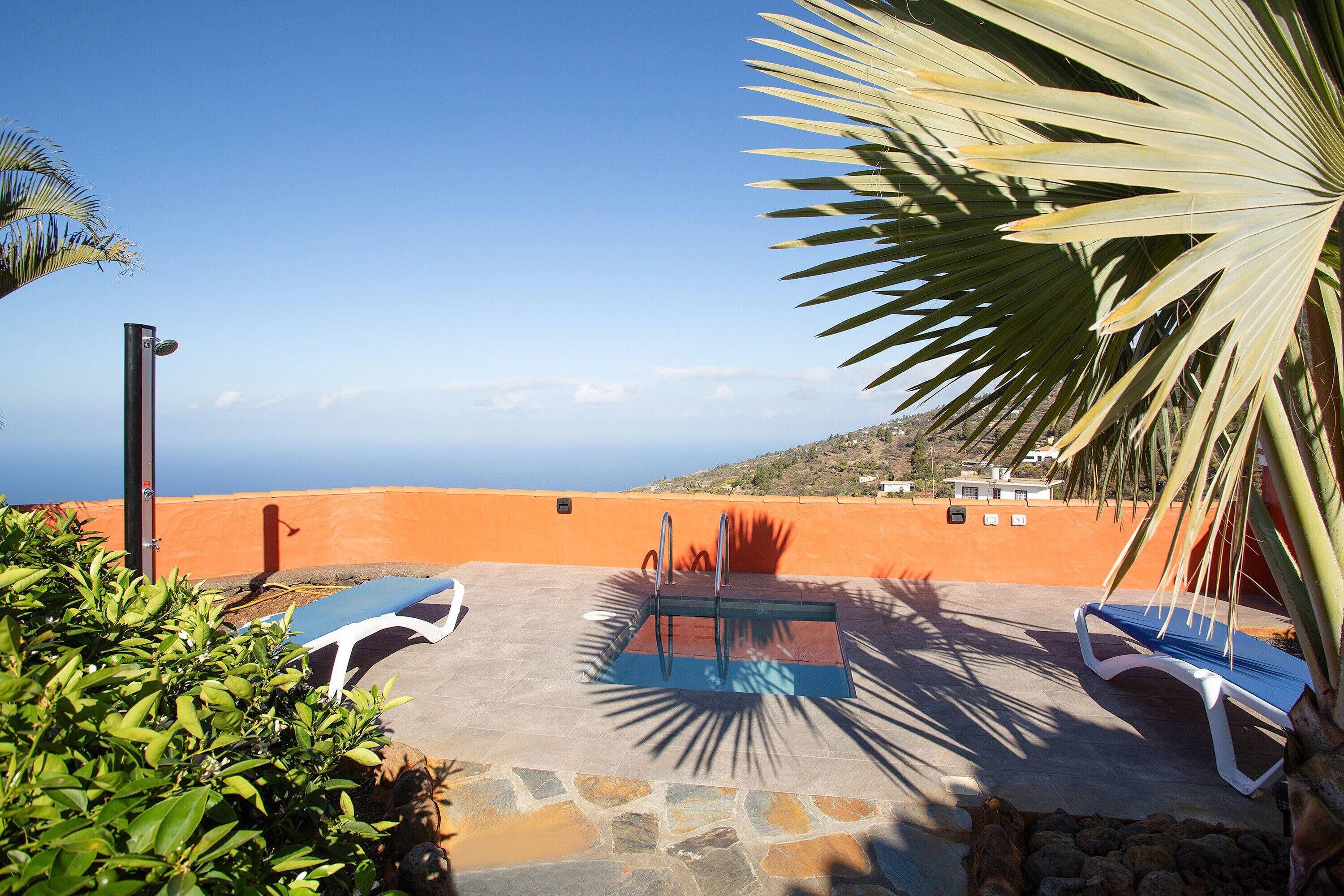 A pool area with lounge chairs and a diving board, surrounded by greenery and overlooking a coastal landscape.