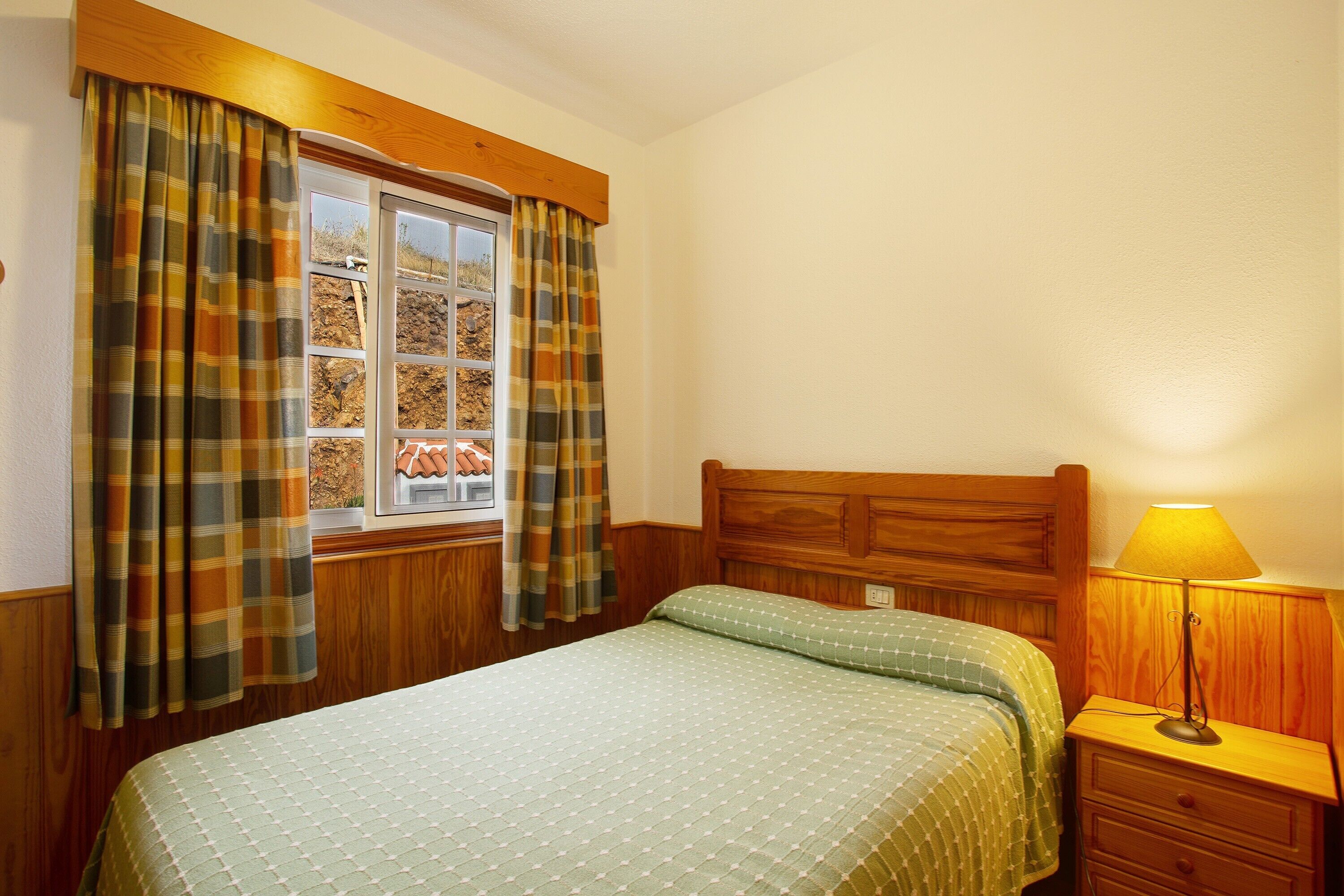A bedroom with a wooden headboard, a window with curtains, a bedside lamp, and a bed with a patterned bedspread.