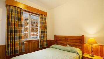 A bedroom with a wooden headboard, a window with curtains, a bedside lamp, and a bed with a patterned bedspread.
