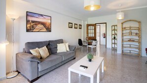 A living room with a sofa, a coffee table, a lamp, a dining area with chairs, and a wall-mounted shelf. - Apartment 'Golden Rana' with Shared Pool, Wi-Fi and Air Conditioning (Albufeira)