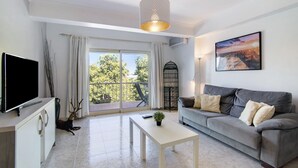 A modern living room with a flat-screen TV, a grey sofa, a white coffee table, and a large window with a view of trees. - Apartment 'Golden Rana' with Shared Pool, Wi-Fi and Air Conditioning (Albufeira)