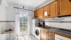 A kitchen with wooden cabinets, a washing machine, a coffee maker, and a window with curtains. - Apartment 'Golden Rana' with Shared Pool, Wi-Fi and Air Conditioning (Albufeira)