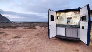 A caravan with an open door, revealing a bed with patterned bedding, a small table, and a window. - Adventure Campingvan with Sea Views and Air Conditioning (Lanzarote)