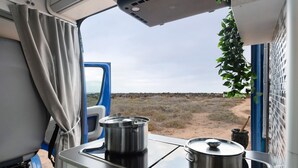 A kitchenette with two pots on a stove, a window with curtains, and a view of a barren landscape. - Adventure Campingvan with Sea Views and Air Conditioning (Lanzarote)