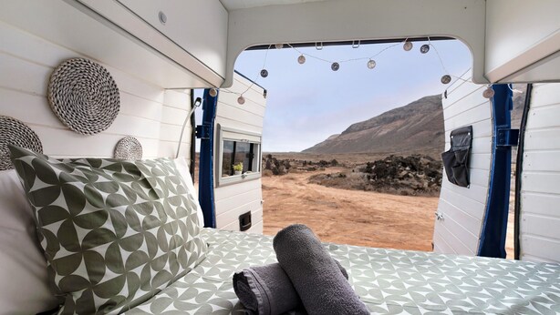 A bed with patterned bedding and a rolled towel, a window with a view of a desert landscape, and decorative wall hangings. - Adventure Campingvan with Sea Views and Air Conditioning (Lanzarote)