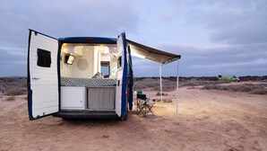 A caravan with an open door, a small kitchenette, a foldable table, and a chair. - Adventure Campingvan with Sea Views and Air Conditioning (Lanzarote)