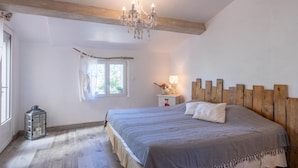 Ein Schlafzimmer mit einem hölzernen Kopfteil, einem Bett mit grauer Bettdecke, einem Nachttisch mit Lampe, einem Fenster mit Vorhängen und einem Laternen auf dem Boden.