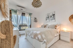 Une chambre à coucher comprenant un lit, des tables de chevet, une fenêtre avec des rideaux, des œuvres d’art murales et un luminaire suspendu.