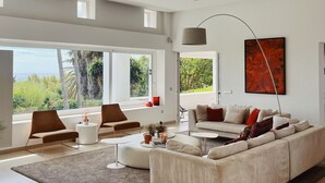 A spacious living room with a large white sofa, a coffee table, and a view of greenery through large windows. - Villa 'Nagueles' with Sea View, Wi-Fi and Air Conditioning (Marbella)