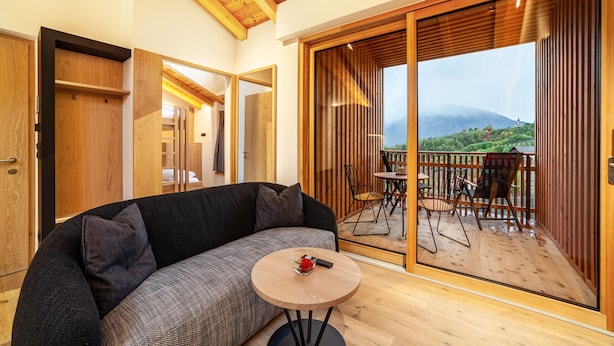 Ein modernes Wohnzimmer mit einer Couch, einem kleinen runden Tisch und einer Schiebetür zu einer Terrasse im Freien mit Blick auf eine gebirgige Landschaft.