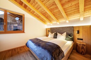 A bedroom with a wooden ceiling, a large bed with white bedding, a window with a view, and a small wooden stool.