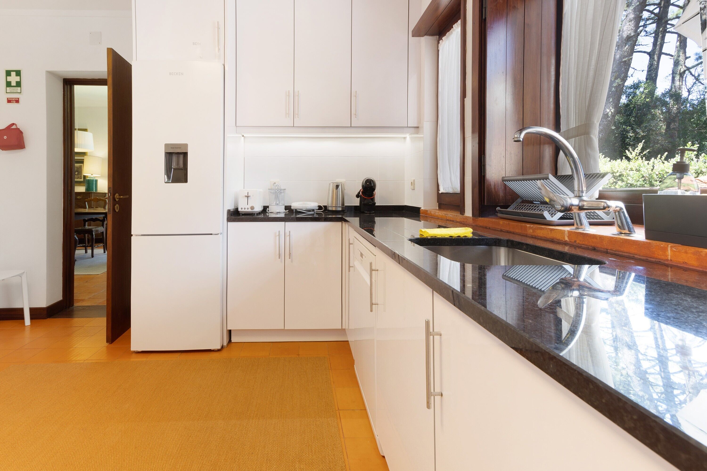 Una cocina moderna con armarios blancos, un lavamanos de acero inoxidable y un amplio ventanal con vistas a árboles.