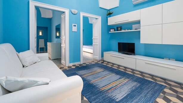 Una sala de estar moderna con un sofá blanco, una pared azul decorativa, un tapete estampado y un televisor de pantalla plana sobre un mueble blanco.