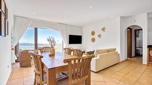 Un área de comedor con una mesa y sillas de madera, un sofá, un televisor de pantalla plana y vistas a través de un ventanal grande.