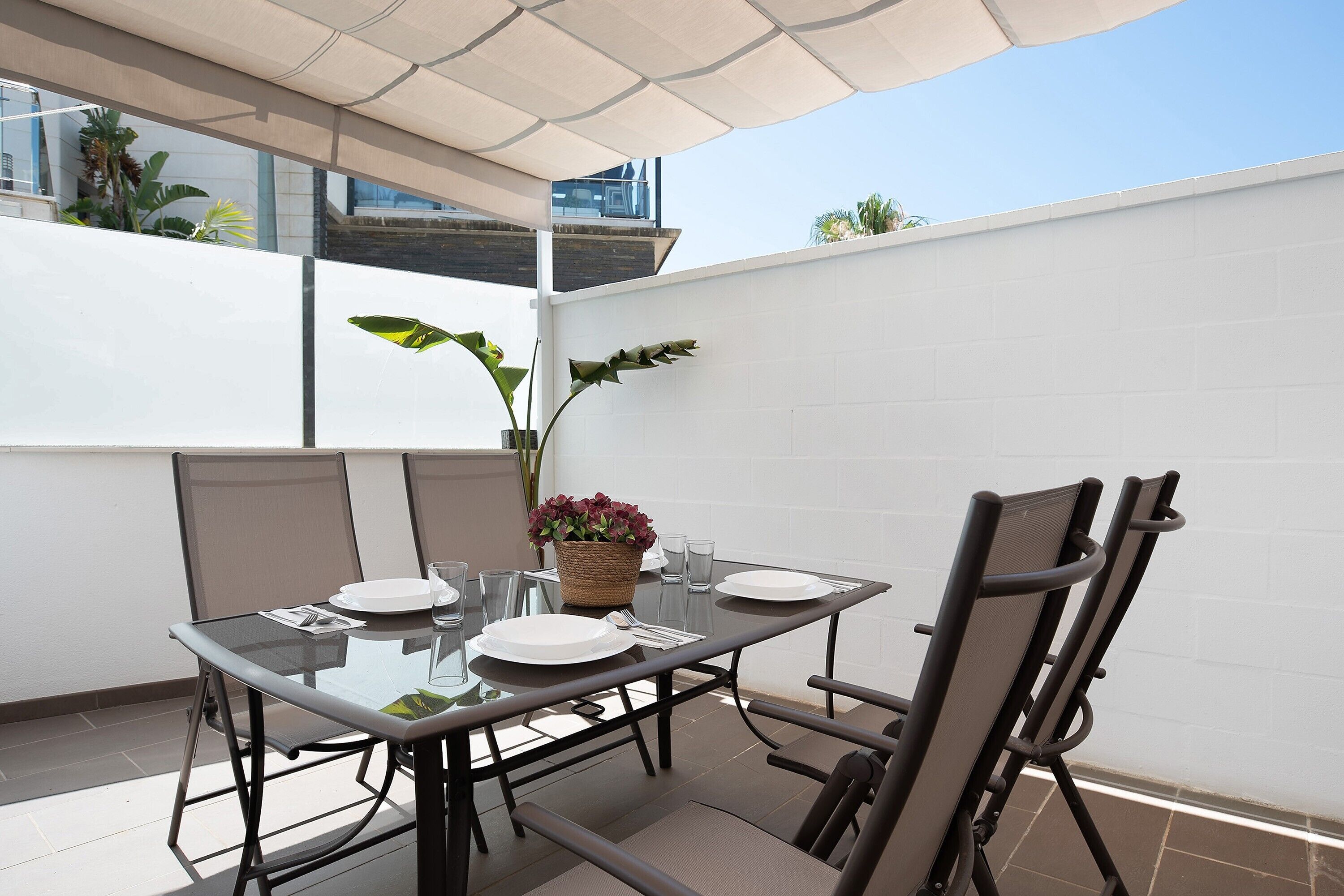 Una mesa con cubierta de vidrio, sillas, un cesto de flores y vista a un cielo despejado.