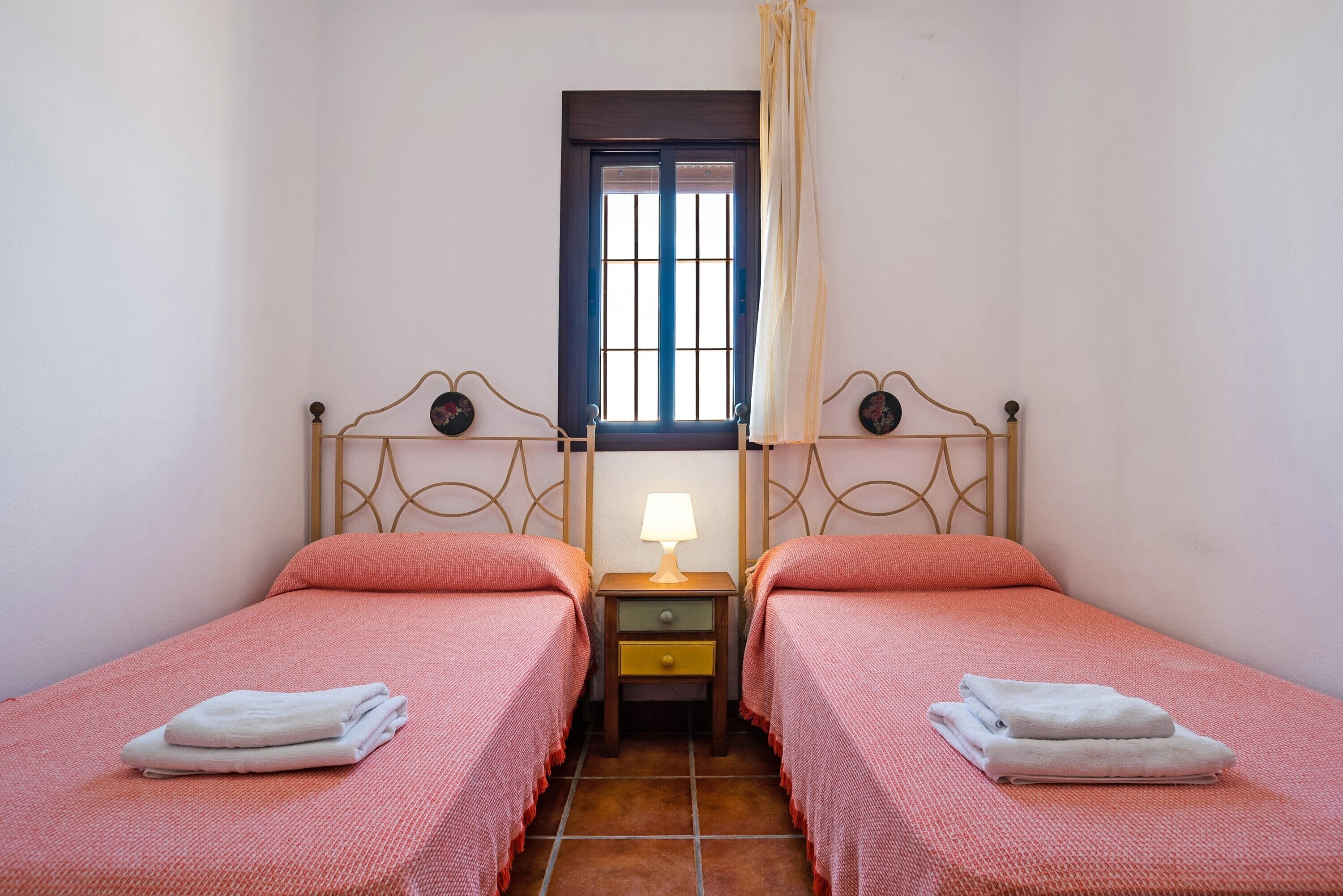 A room with two beds, a window with curtains, a nightstand with a lamp, and a small drawer.