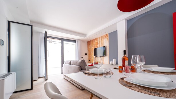 A modern dining area with a long table set for two, a flat-screen TV mounted on the wall, and a sofa in the background.