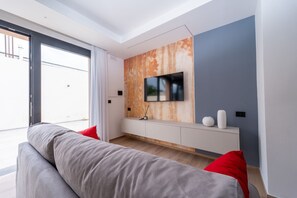 Un salon moderne avec un canapé gris, des coussins rouges, un téléviseur fixé sur un mur à texture, et une console blanche.