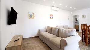 Un salon avec un canapé beige, une table d’appoint en bois, un téléviseur fixé au mur et un coin repas avec des chaises en bois.