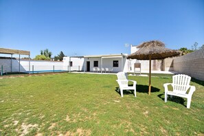 Un espacio al aire libre con césped, sillas blancas y un paraguas de paja, un edificio blanco con ventanas y un muro de ladrillo.