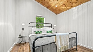 A bedroom with a bed, bedside lamps, a small table, and a window with a view of greenery.