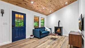 A living room with a blue door, two windows with views of greenery, a blue sofa, a fireplace, and a wooden ceiling.