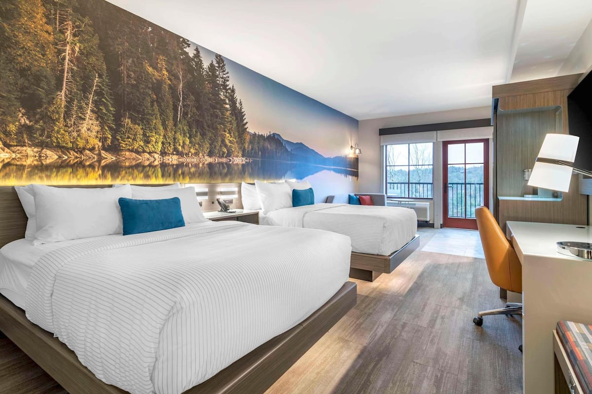A hotel room with two beds, a desk, a chair, and a large forest mural on the wall.