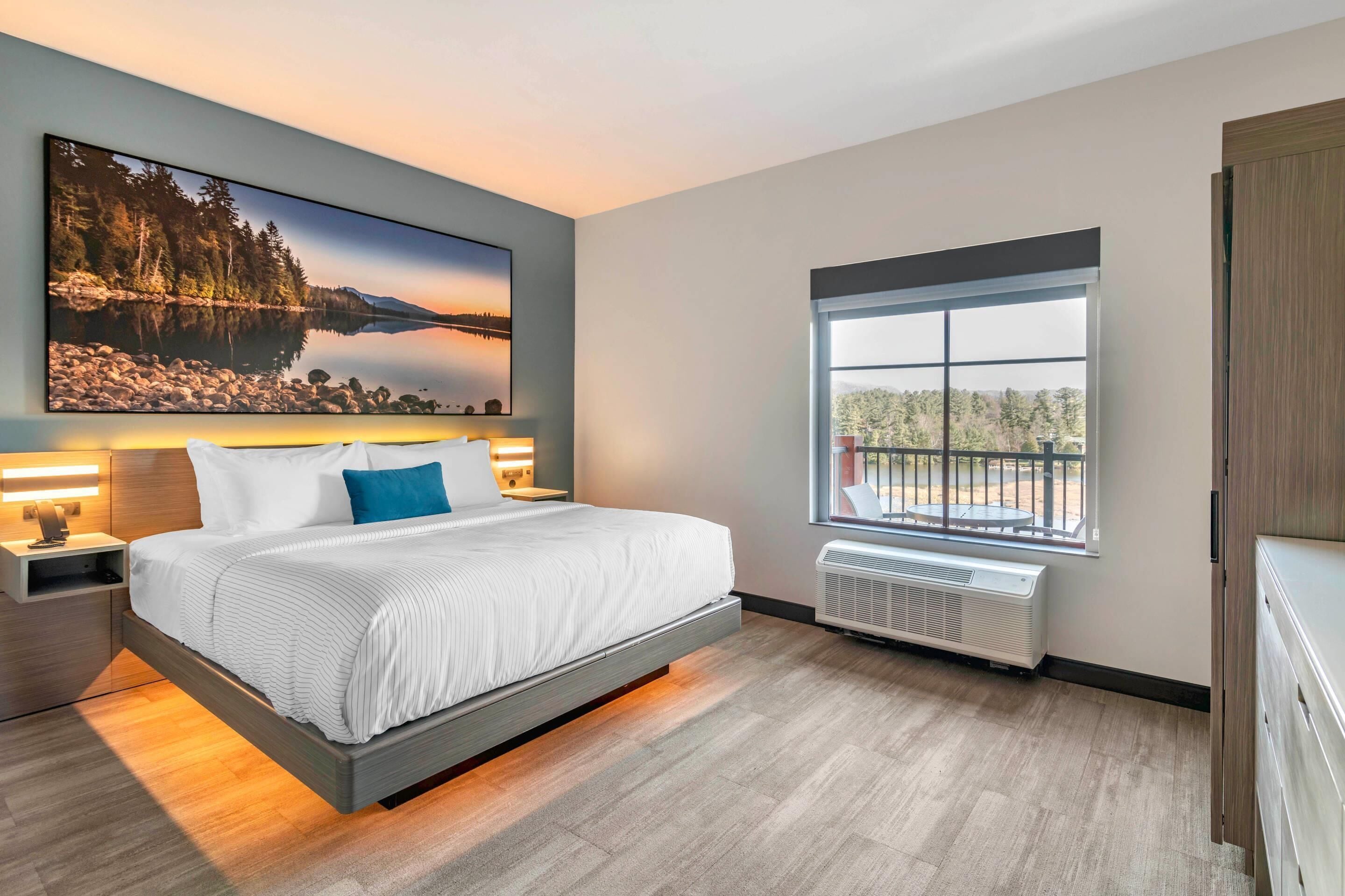 A modern bedroom with a large bed, a nightstand, a wall-mounted TV, and a window with a view of trees and a building.