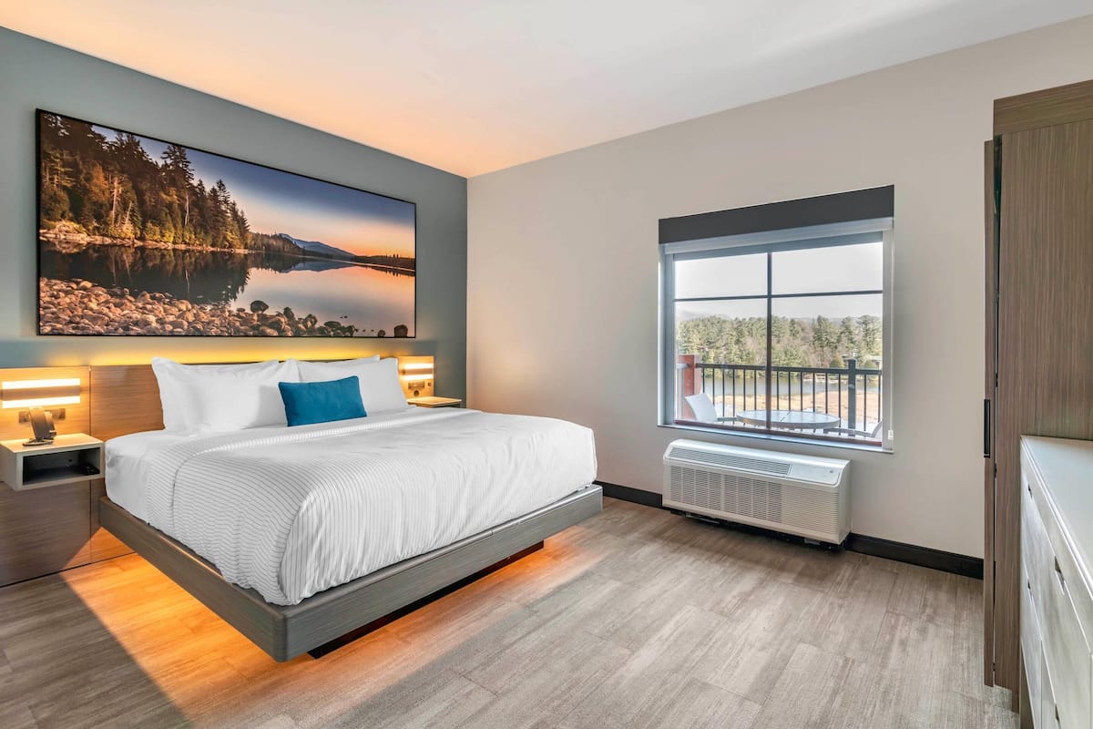 A modern bedroom with a large bed, a nightstand, a wall-mounted TV, and a window with a view of trees and a building.