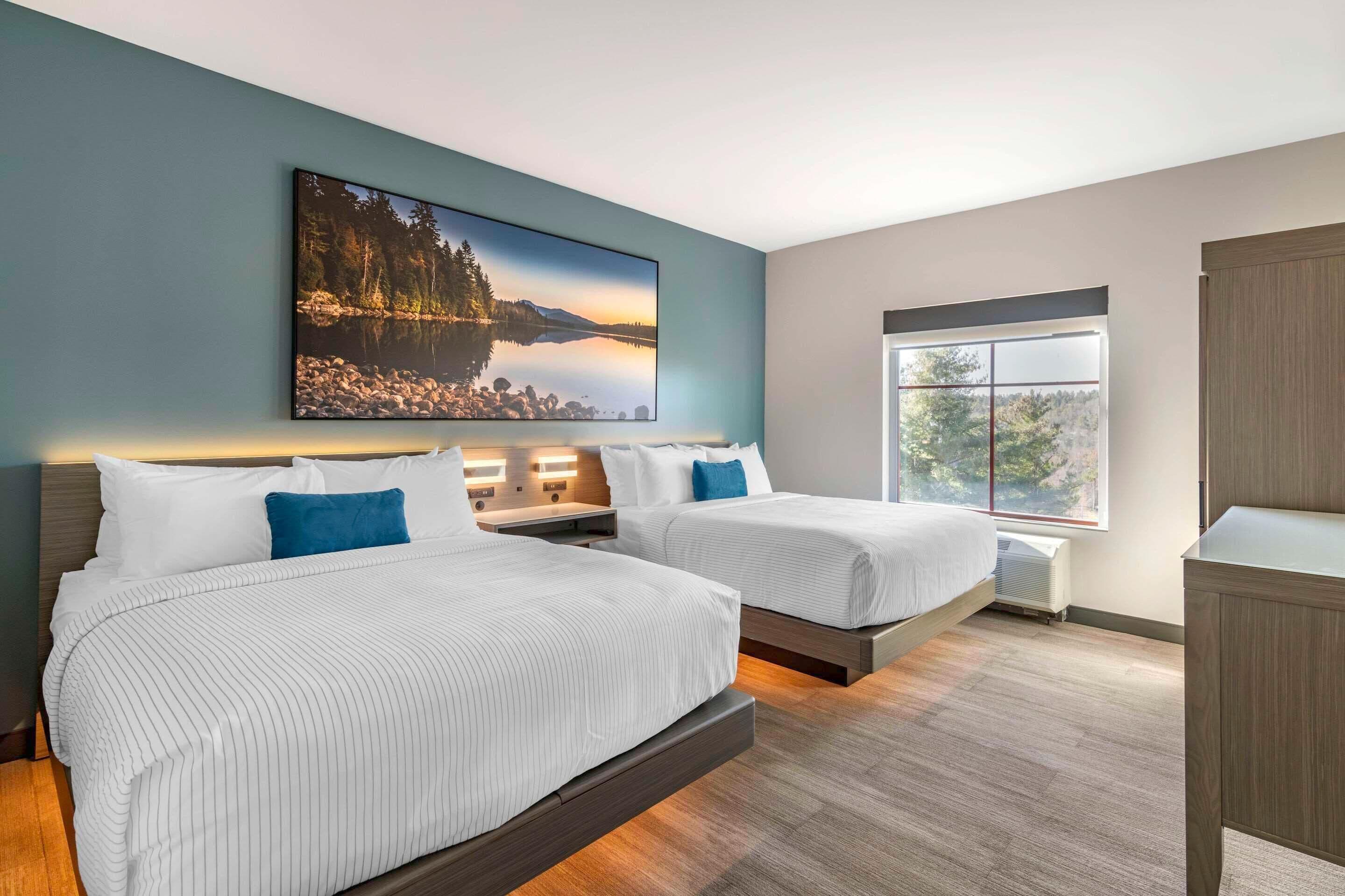 A modern hotel room with two beds, a large window, and a scenic painting on the wall.