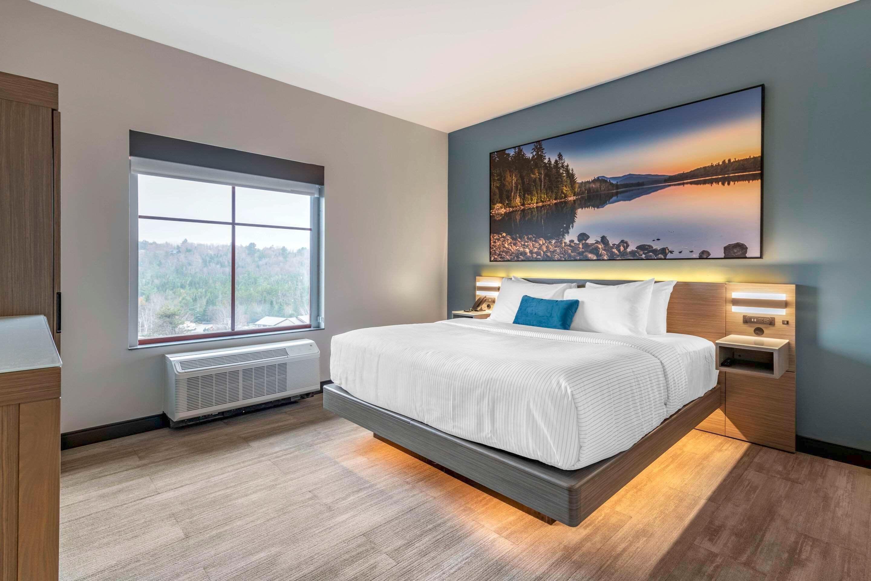 A modern bedroom with a large bed, a window with a view, a mounted painting, and a bedside table with a lamp.