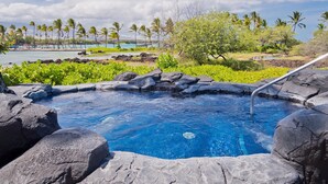 A hot spring surrounded by rocks and palm trees, with a view of the beach and ocean in the background.