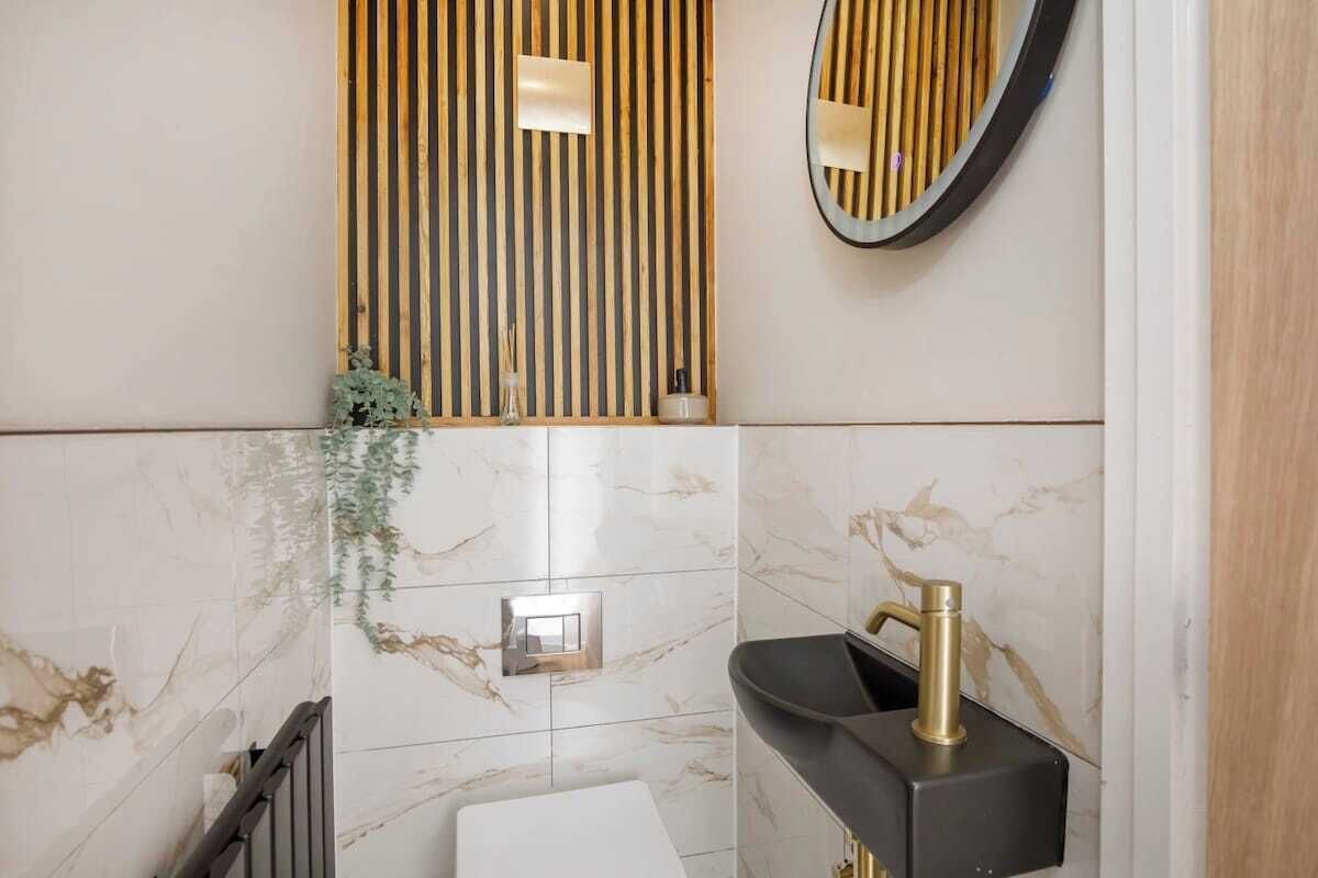 Une salle de bain avec un mur en marbre, un panneau mural en bois à lattes, un miroir rond, un lavabo noir et des toilettes blanches.