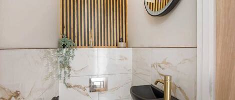 Une salle de bain avec un mur en marbre, un panneau mural en bois à lattes, un miroir rond, un lavabo noir et des toilettes blanches.