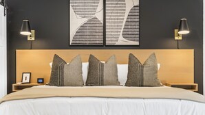 A modern hotel room with a large bed, three pillows, two bedside tables, a nightstand, and wall art.
