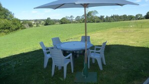 A round table with white plastic chairs and a large umbrella in a grassy outdoor area. - Gîte Indépendant Dans Maison Propriétaires (Saint-Laurent-en-Grandvaux)