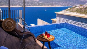 Uma área à beira da piscina com vista para o mar, uma espreguiçadeira, um guarda-sol e uma mesa pequena com bebidas.