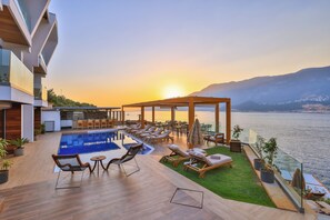 A modern outdoor pool area with lounge chairs and a pergola overlooking a body of water and mountains.