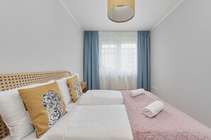 Ett hotellrum med två sängar, ett fönster med blå gardiner, en vit sänggavel och en väggmonterad lampa.