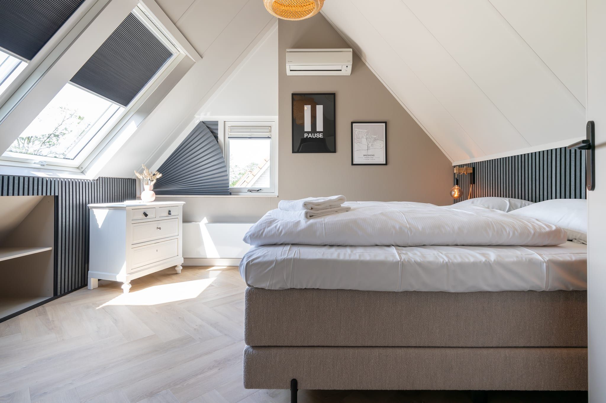 A modern bedroom with a sloped ceiling, a bed, a nightstand, a wall-mounted air conditioner, and a window with blinds.