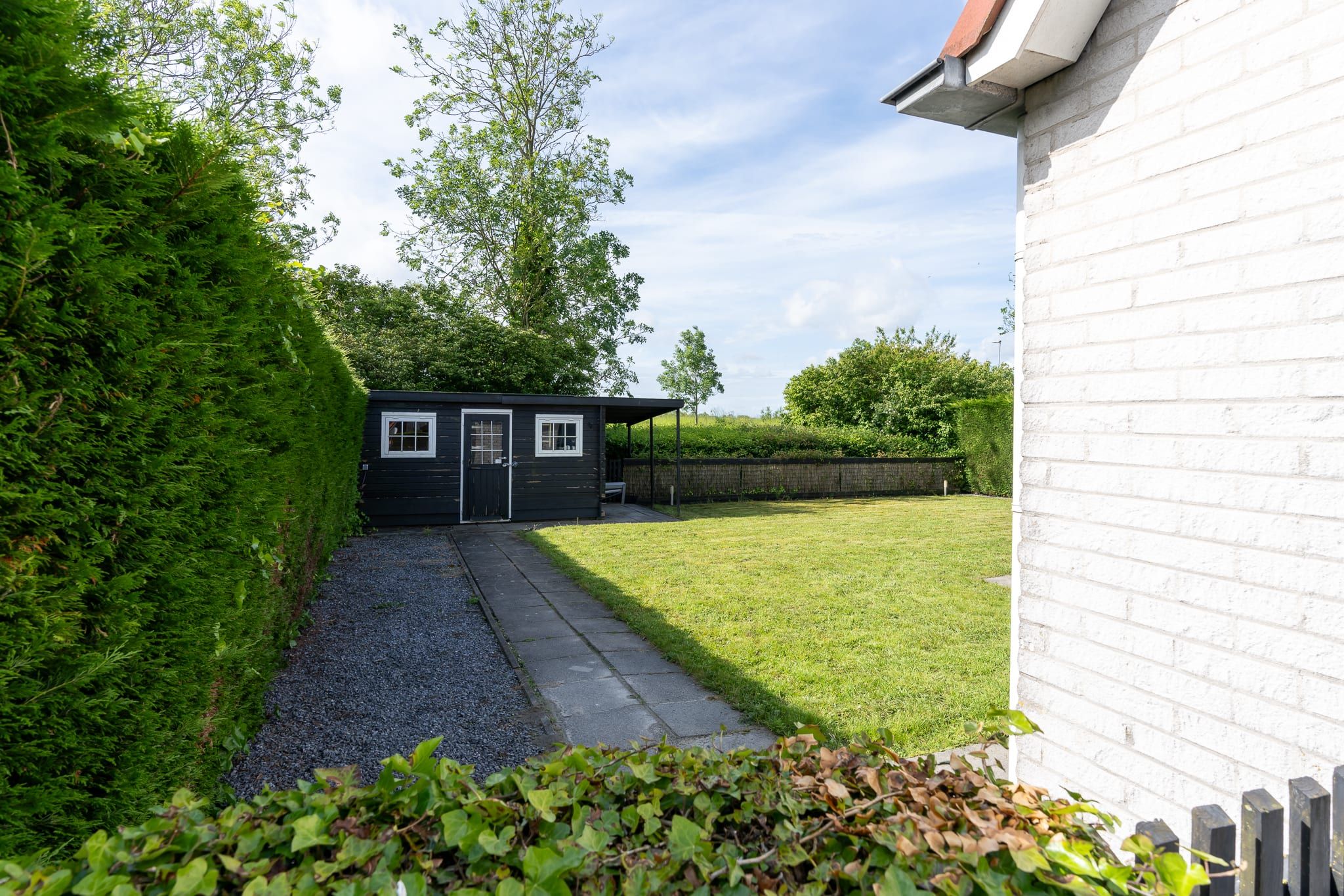 Ein kleiner, dunkel gefärbter Schuppen mit Tür und Fenstern, welcher in einem grasbewachsenen Hof mit Kiesweg steht.
